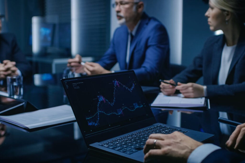 Business professionals in formal attire sit around a conference table, discussing data. A laptop displaying a financial graph with fluctuating lines is open in the foreground. Papers and notebooks are also visible on the table.