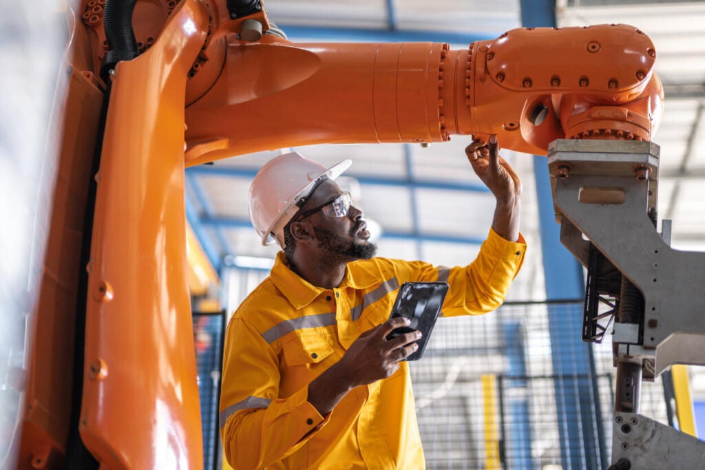 A worker in a yellow safety jacket and white hard hat inspects a large orange industrial robot arm while holding a tablet in a factory setting.