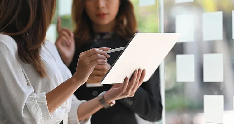 Deux femmes se tiennent à l'intérieur&nbsp;; l'une tient une tablette numérique tandis que l'autre fait un geste du doigt. Toutes deux sont concentrées sur l'écran, semblant engagées dans une discussion ou une collaboration.