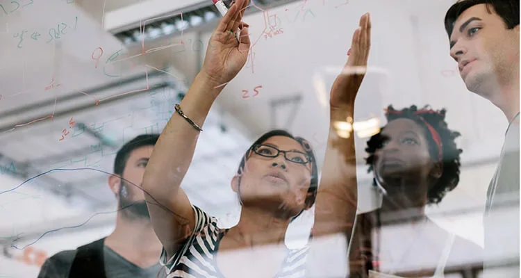Four people stand behind a transparent board covered in graphs and equations; one person gestures towards the board, explaining something to the group.
