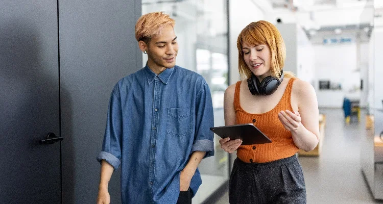 Two people walk together indoors. One is holding a tablet and wearing headphones around their neck, while the other smiles and looks at the tablet. They appear to be having a casual conversation in a modern office setting.