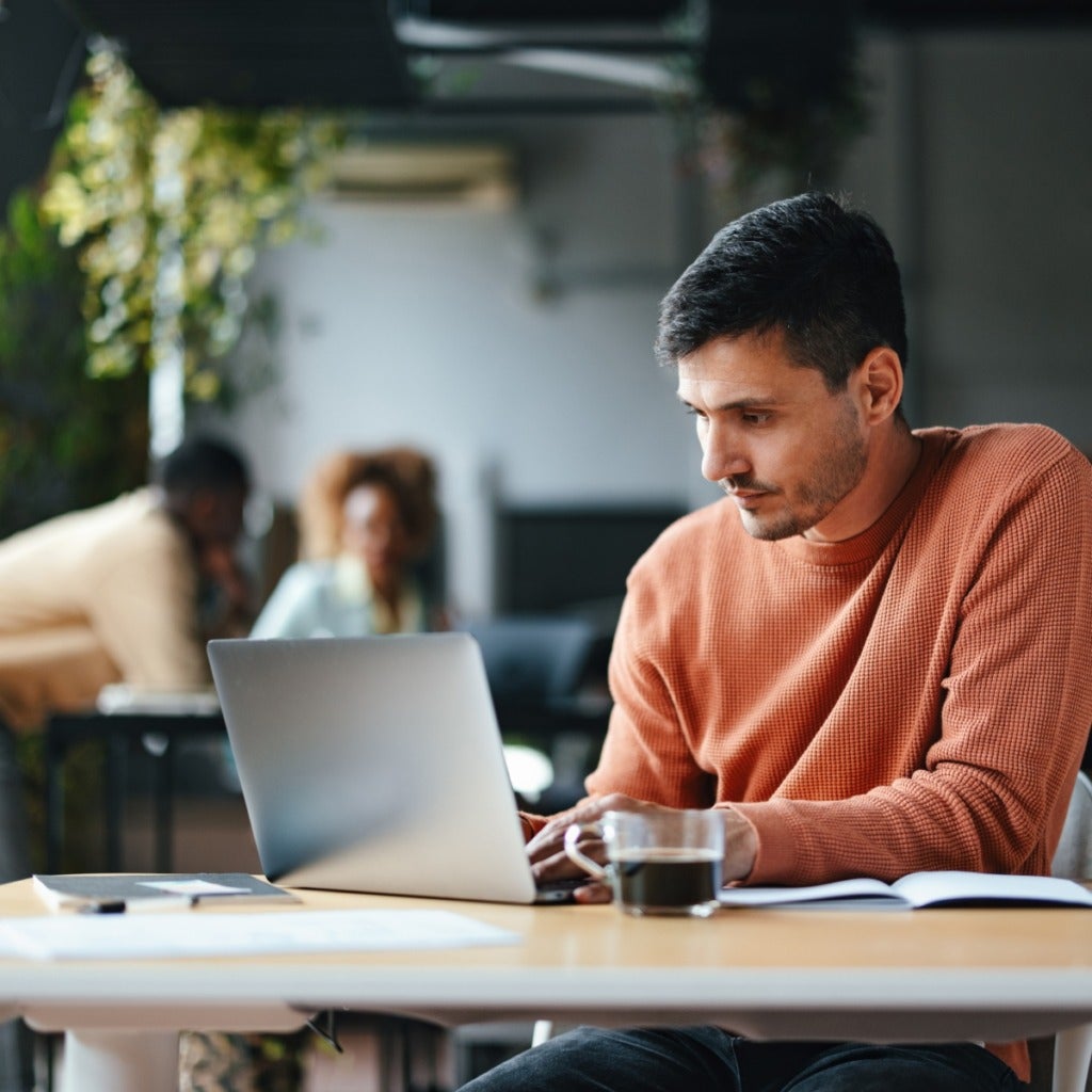 A man in an orange sweater works on a laptop at a desk with a notebook and coffee cup; two people talk in the blurred background of a modern, well-lit workspace with plants.