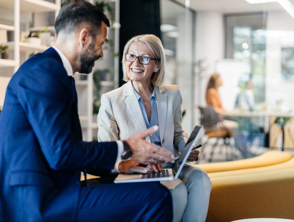 Two business professionals, a man and a woman, are sitting and talking in a modern office. The woman is smiling and holding a laptop, while the man gestures as they have a discussion.