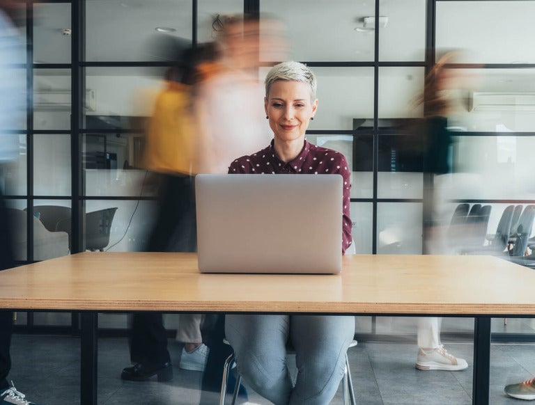 A woman working on a laptop, with co-workers walking behind her.